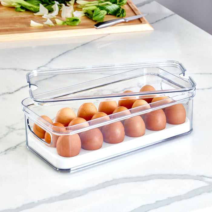 The Stackable Fridge Egg Bin from iDesign, made with recycled plastic, holds a dozen brown eggs on a white surface. A transparent lid reveals the eggs while a wooden cutting board and chopped vegetables on the marble counter suggest culinary possibilities.