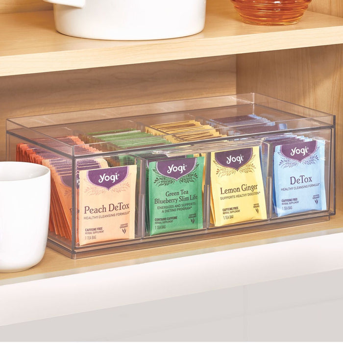 An iDesign Stackable Tea Organizer, crafted from recycled plastic and resting on a wooden shelf, holds various Yogi tea packets—Peach DeTox, Green Tea Blueberry Slim Life, Lemon Ginger, and DeTox. A partially visible white mug is on the left.