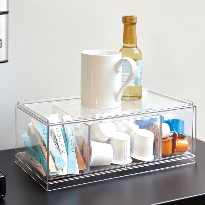 A white mug and a small bottle sit on an iDesign Stackable Tea Organizer made with recycled plastic, ideal for tea packets and coffee pods. Its handy compartments rest elegantly on a dark surface against a light-colored wall.