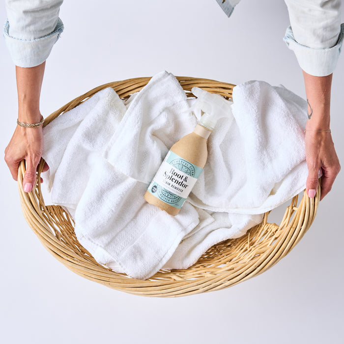 A person holds a wicker laundry basket with white towels and a brown spray bottle labeled Stain Remover by Root & Splendor. Made with natural ingredients, this eco-friendly packaging enhances sustainability against the simple white background.