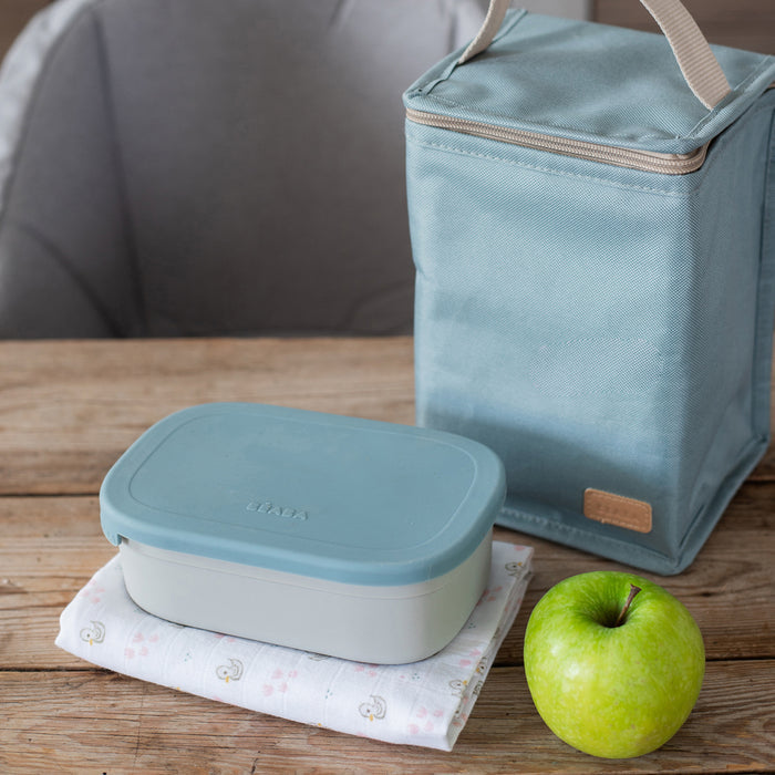 A blue, eco-friendly BEABA Stainless Steel Bento Lunch Box and matching insulated bag rest on a wooden table. A green apple and patterned cloth enhance the scene against a gray chair, ideal for those who value style and sustainability in their daily routine.