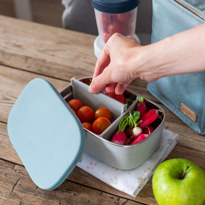 A hand reaches into an eco-friendly container with cherry tomatoes and radishes on a wooden table. Nearby are a green apple and blue insulated lunch bag, a smoothie cup, and the BEABA Stainless Steel Bento Lunch Box with its lid open.