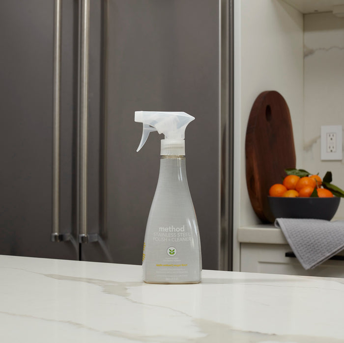 A bottle of method Stainless Steel Cleaner + Polish sits on a white kitchen countertop. In the background, a stainless steel refrigerator gleams, and a bowl of oranges on a cutting board near a gray towel reflects the surface-safe polish sprays brilliance.
