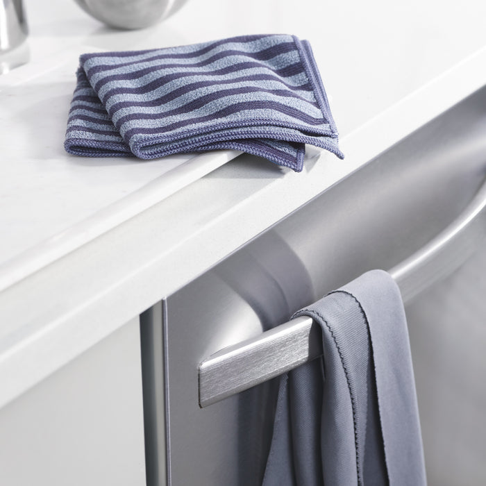 A folded blue-striped cloth lies on a white countertop beside a gray towel hanging from the dishwasher handle, ready for use with your E-Cloth Stainless Steel Cleaning Kit.