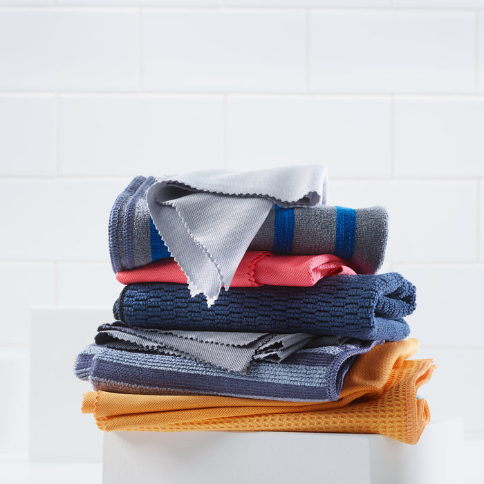 Against a white tiled background, colorful folded towels in orange, blue, pink, and gray with zigzag edges are neatly arranged to complement the sleek aesthetic of the E-Cloth Stainless Steel Cleaning Kit in this beautifully organized space.