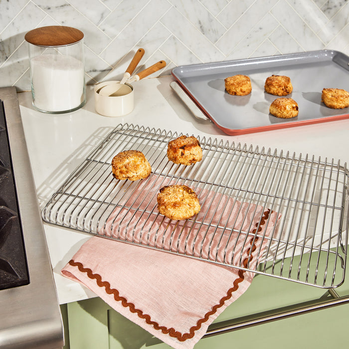 Freshly baked scones rest on a Caraway Stainless Steel Cooling Rack, highlighted by a pink cloth, next to a baking tray filled with oven-ready treats. Nearby, a sugar jar, stainless steel utensils, and a small cream pitcher adorn the white kitchen counter with its tiled backsplash.