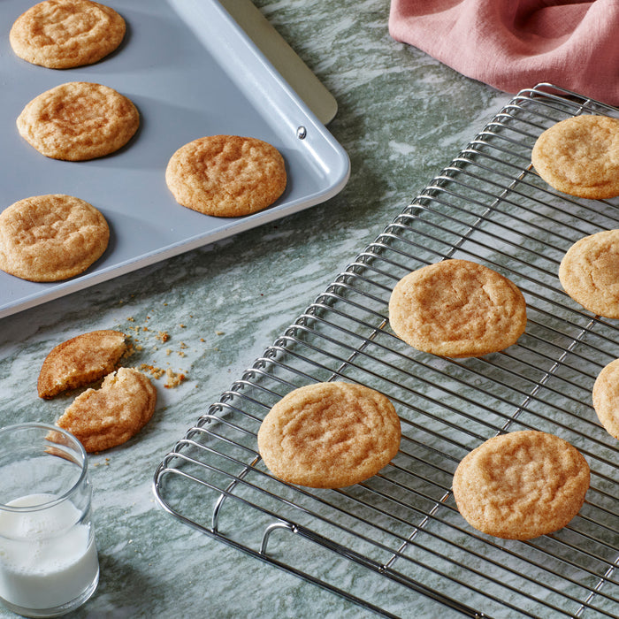 A batch of golden-brown cookies sits on a Caraway Stainless Steel Cooling Rack beside an oven-safe baking sheet filled with more treats. One cookie is broken on a marble surface near a glass of milk, with a pink cloth peeking from the corner.