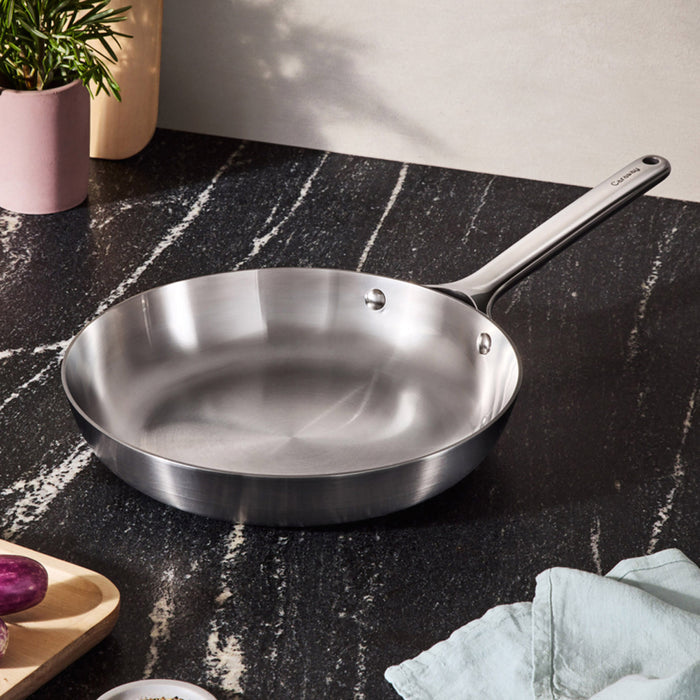 On a dark marble countertop, the Caraway 8 Stainless Steel Fry Pan, featuring non-toxic 5-ply construction and a long handle, is showcased. Nearby are a cutting board with purple onion and garlic, a light blue cloth, and a pink plant pot, all bathed in natural light.