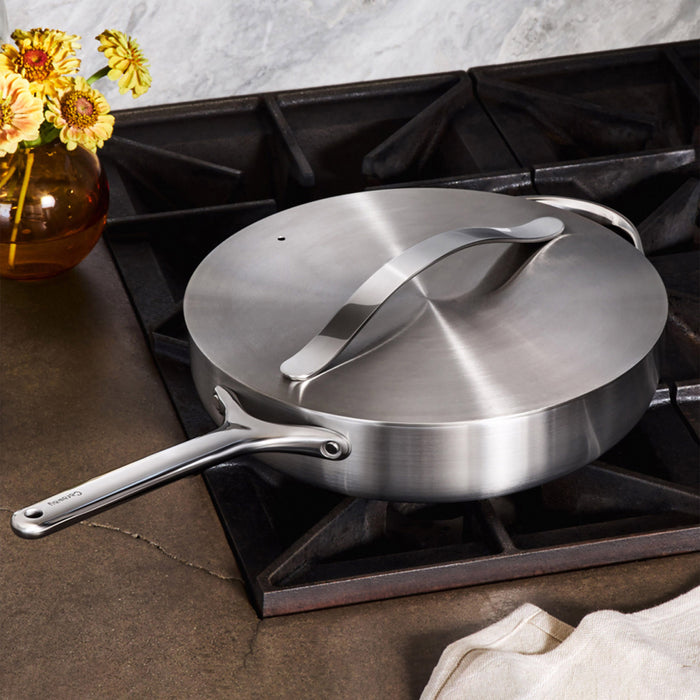 The Caraway Stainless Steel Sauté Pan, 4.5 qt, with non-toxic 5-ply construction and a long handle in a sleek design is on the stove. A small vase with yellow flowers sits to the left, and a folded cloth lies at the bottom right on the dark countertop.