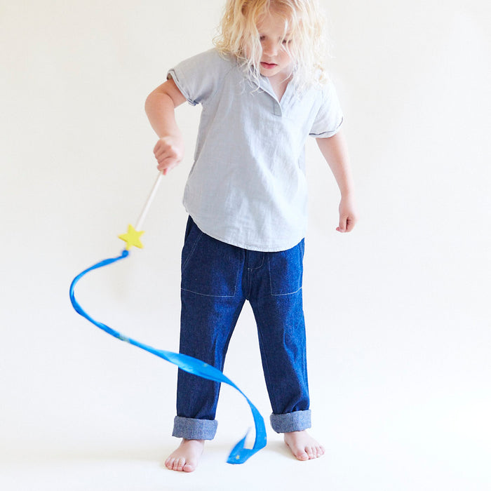 A child with long blond hair, wearing a light blue shirt and rolled-up jeans, joyfully engages in open-ended play. Barefoot against a white backdrop, they twirl the Star Streamer by Sarahs Silks—a blue ribbon wand with a yellow star—enhancing gross motor skills while creating colorful patterns.