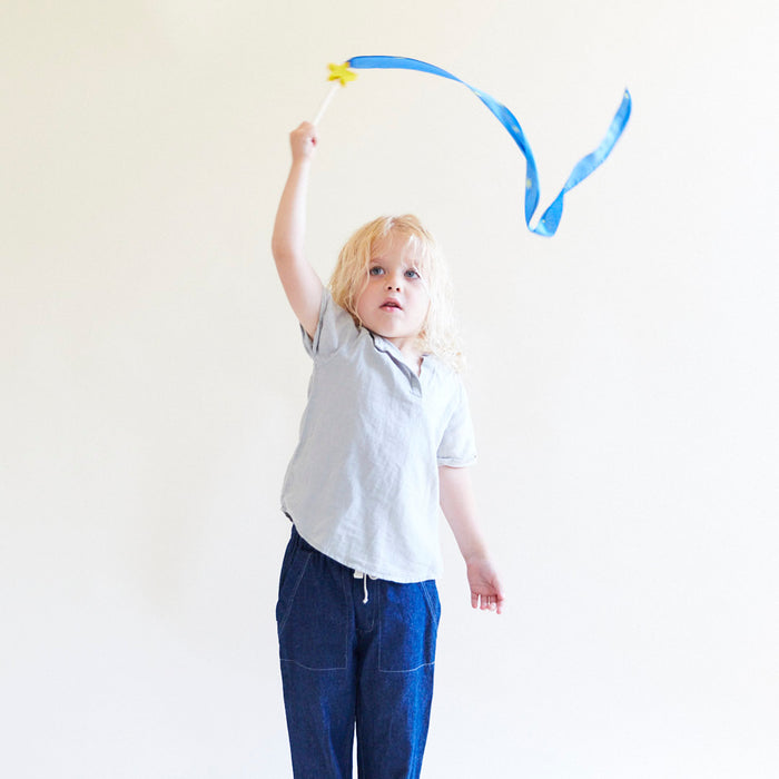A young child with long blonde hair, dressed in a light gray shirt and dark pants, waves Sarahs Silks Star Streamer in the air. The blue and yellow ribbon fosters open-ended play and enhances gross motor skills against a plain light background.