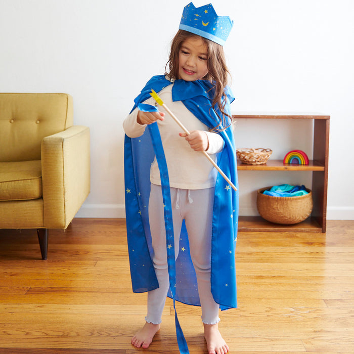 A child engaged in open-ended play wears a wizard outfit with a blue cape and crown adorned with stars. They hold Sarahs Silks Star Streamer wand and stand on a wooden floor in a room with a yellow armchair, shelf, and toys that foster gross motor skill development.