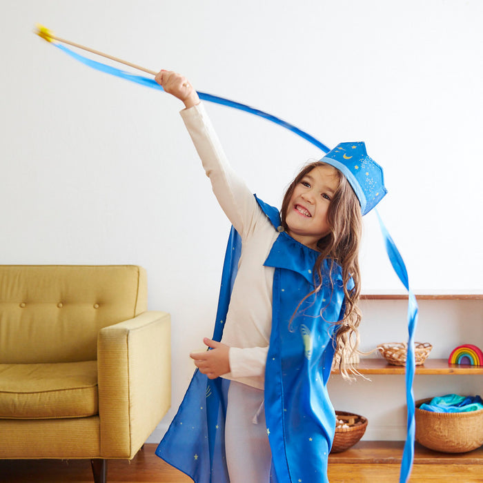In the living room, a child joyfully waves a Star Streamer wand by Sarahs Silks in a blue wizard costume adorned with stars, fostering gross motor skills. A yellow chair and colorful toys in the background invite open-ended play.