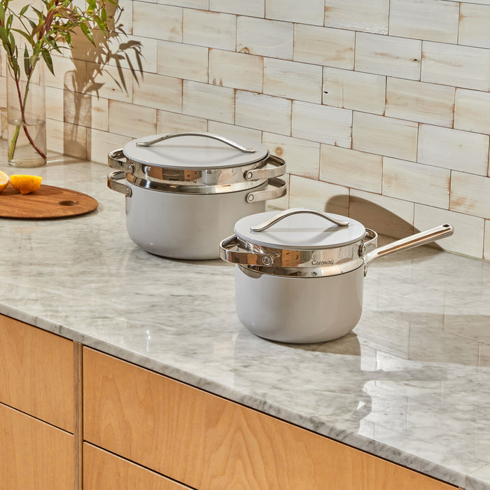 The Caraway Steamer Duo, two white pots with lids, sits on a marble countertop, displaying its sleek non-toxic ceramic coating. A wooden cabinet below complements the tiled wall background, enhanced by a small wooden board with sliced lemon adding freshness to this charming kitchen scene.