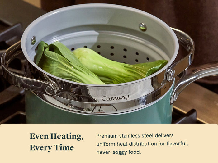 A green Caraway Steamer Duo sits on a stove with leafy bok choy steaming inside its stainless steel basket. Text below reads: Even Heating, Every Time. The non-toxic Steamer Duo ensures flavorful, never-soggy food.