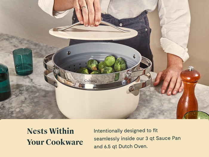 A person lifts the lid off a Caraway Steamer Duo, revealing Brussels sprouts in its stainless steel basket. The pot rests on a marble counter beside turquoise glasses and a wooden pepper grinder. Text highlights the nesting design.
