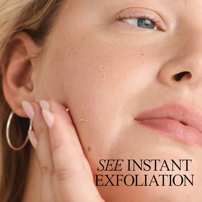 Close-up of a woman's face with a focus on her cheek being gently exfoliated, showing small bits of exfoliant on her skin. She has hoop earrings, and her light pink nails are visible. Text on the image reads, "SEE INSTANT EXFOLIATION.
