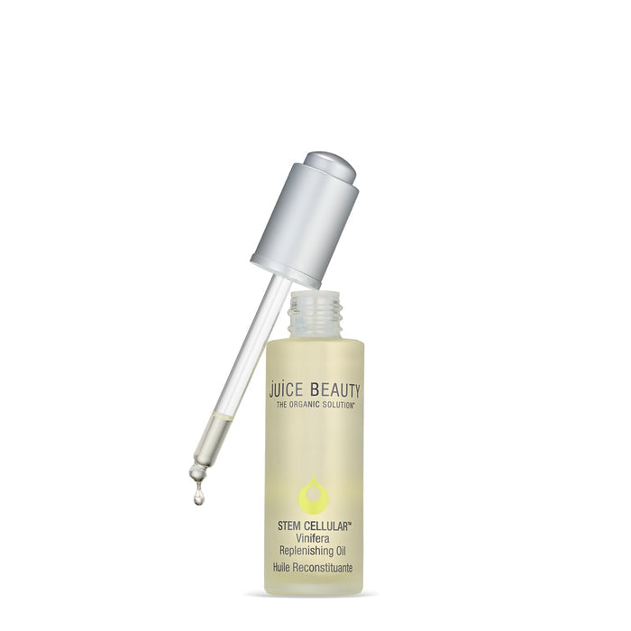 A clear dropper with liquid is suspended above a translucent bottle labeled Juice Beautys Stem Cellular Vinifera Replenishing Oil + Primer. The sleek silver cap complements the falling drop from the dropper, set against a pristine white background.