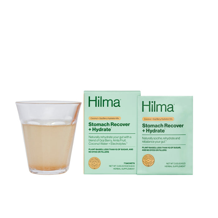 A light-colored liquid in a glass sits next to two boxes of Hilmas Stomach Recover + Hydrate. The green packaging promotes its natural ingredients, with gastroprotective herbs for gut health and essential electrolytes for an ideal stomach reset.