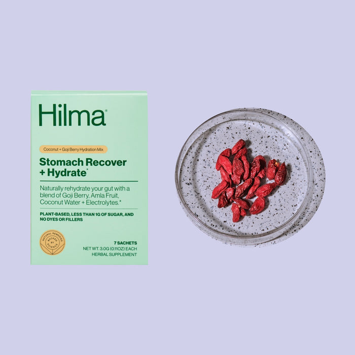 A box of Hilmas Stomach Recover + Hydrate, set against a light purple backdrop with a speckled ceramic dish holding dried goji berries, highlights the stomach-resetting benefits of coconut water, goji berry, and amla fruit—packed with electrolytes and gastroprotective herbs.