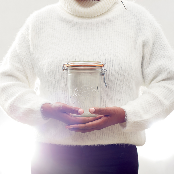 A person in a white sweater holds an empty Le Parfait Storage and Canning Glass Jar, 1L with a clasped lid against a bright white background, highlighting an elegant plastic-free food storage option.