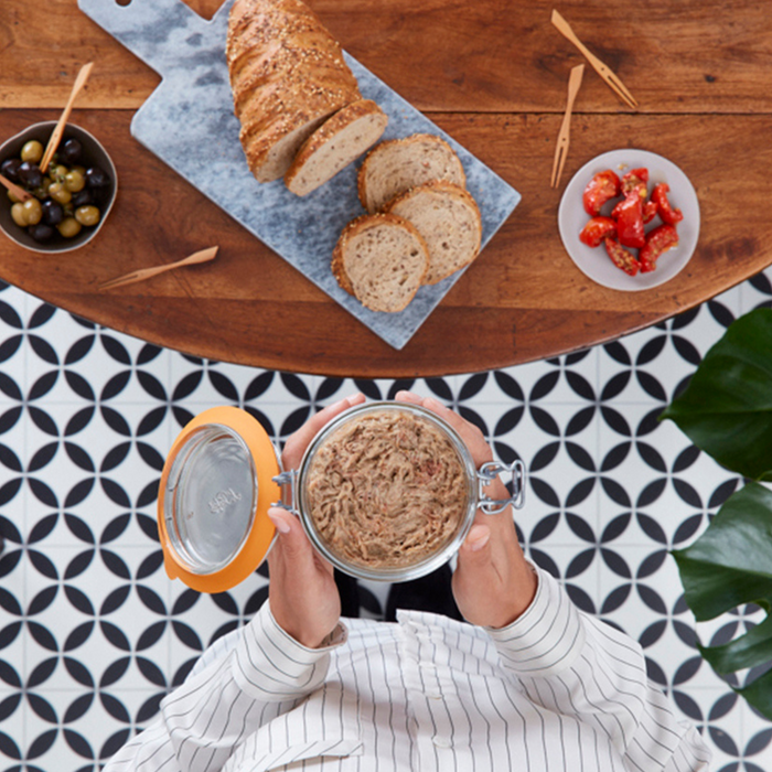 A person in a striped shirt holds a 1L Le Parfait Storage and Canning Glass Jar above a patterned floor, with sliced bread on a cutting board, olives in a bowl, and cherry tomatoes on a plate nearby, enjoying the moment with plastic-free food storage.