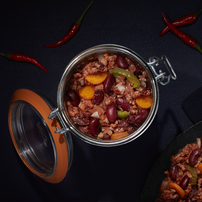 A Le Parfait Storage and Canning Glass Jar, 1L, is filled with chili containing beans, minced meat, carrot slices, and green bell peppers on a dark background. Red chili peppers scattered around make it an ideal showcase of plastic-free food storage.