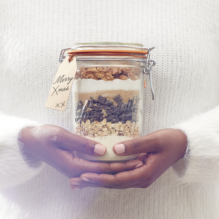 A person in a white sweater holds a Le Parfait Storage and Canning Glass Jar, 1L, filled with baking ingredients like oats and walnuts. The jars tag says Merry Xmas xx, adding a personal holiday touch.