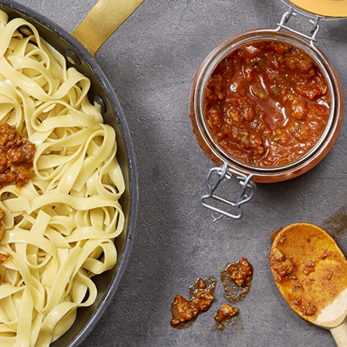 A skillet with cooked tagliatelle pasta is partially covered in a rich red meat sauce. An open 500ml Le Parfait Storage and Canning Glass Jar sits nearby, with a wooden spoon featuring some sauce resting beside the jar on a gray surface.