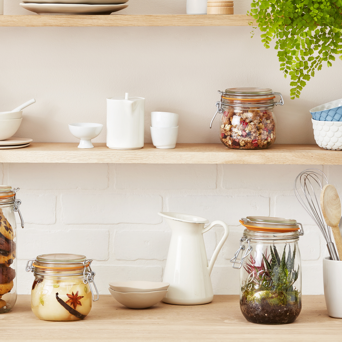 The kitchen shelves feature Le Parfaits 500ml Storage and Canning Glass Jars filled with plants and dried ingredients. White ceramic dishes and pitchers enhance the display, while a green plant adds vibrant color to the upper shelf, all in recyclable glass jars.