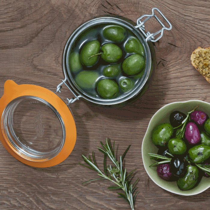 A 500ml Le Parfait Storage and Canning Glass Jar with its recyclable glass and airtight seal is open on a wooden table, next to a green bowl filled with green and black olives. The rustic scene is completed by a sprig of rosemary and crusty bread.