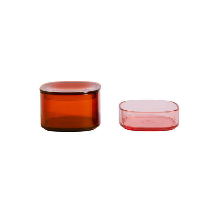 Two stackable Storage Gems from Open Spaces sit on a white background: a taller amber-hued cylinder with a lid and a shorter, soft pink square-shaped piece. These glass containers elegantly blend function and style.
