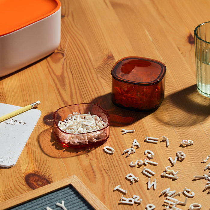The table displays Open Spaces Storage Gems, plastic letters, a glass cup, notebook, and pencil. Sunlight casts warm shadows on the wood surface.