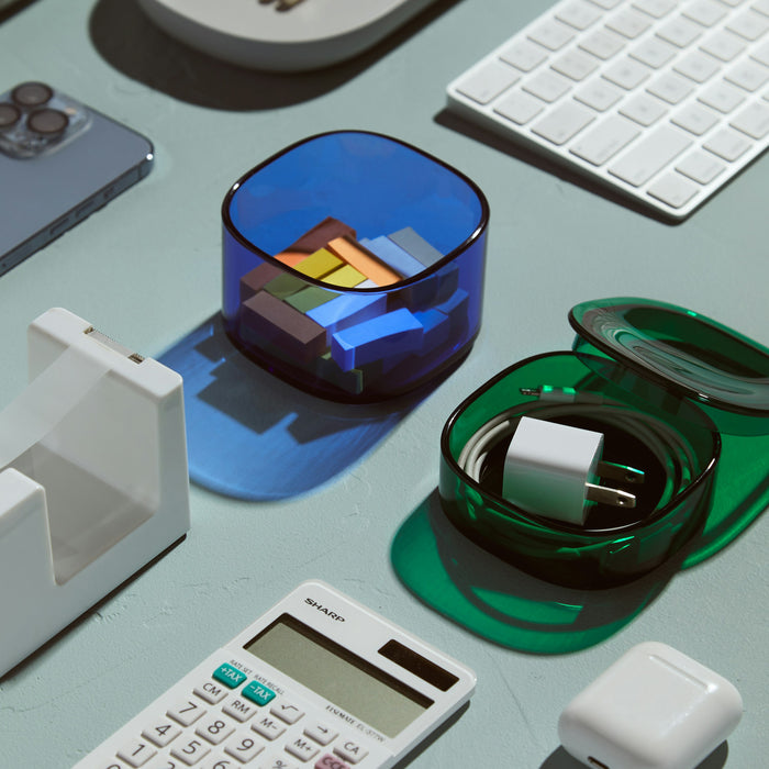 A flat lay of office items on a light green surface features essentials like a smartphone, tape dispenser, calculator, and keyboards. Open Spaces Storage Gems elegantly showcase erasers in blue and a charging cable with plug in green, adding an organized touch to the workspace.
