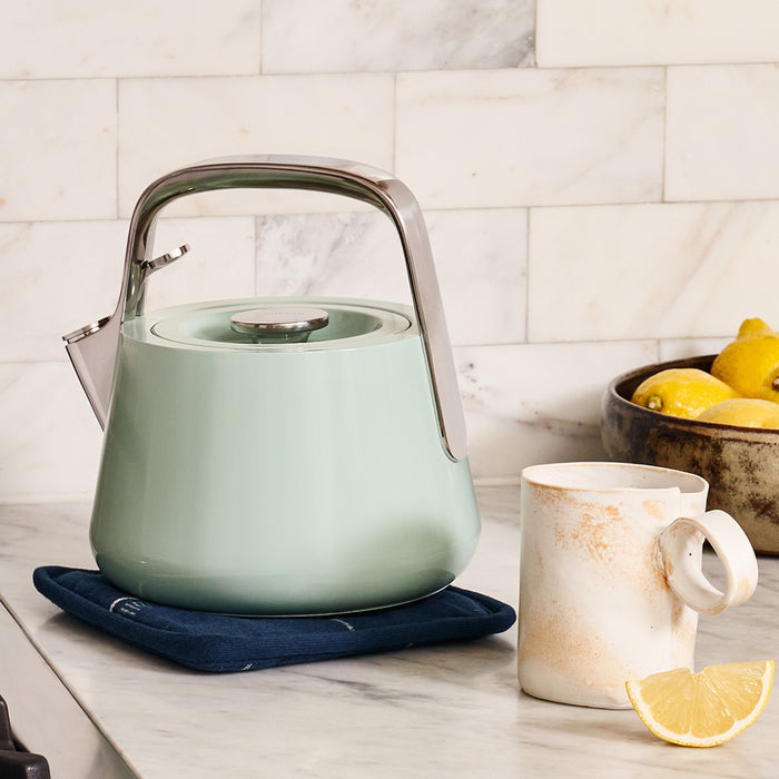 A Caraway Stovetop Whistling Tea Kettle in pastel green with a sleek silver handle sits on a blue mat on a marble countertop, next to a ceramic mug featuring a light marble pattern and a lemon slice. A bowl of lemons is visible in the background.