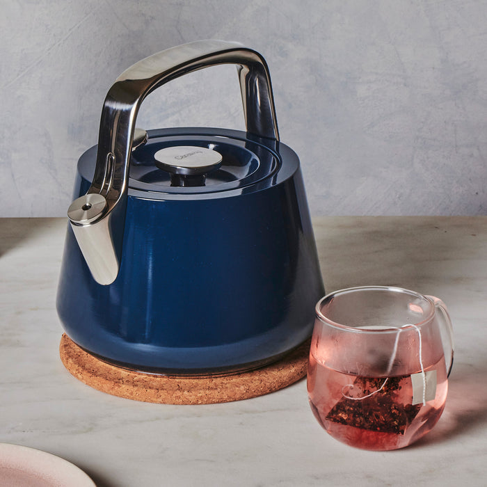 A sleek, modern blue Caraway Stovetop Whistling Tea Kettle rests on a cork coaster. Beside it, a glass mug holds a teabag steeping in pink-tinted water against a textured gray backdrop.