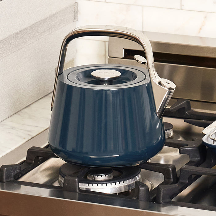 A Caraway Stovetop Whistling Tea Kettle, featuring a blue design and stainless steel handle, is on a gas stove burner in a kitchen with non-toxic materials, marble countertop, and light tile backsplash.
