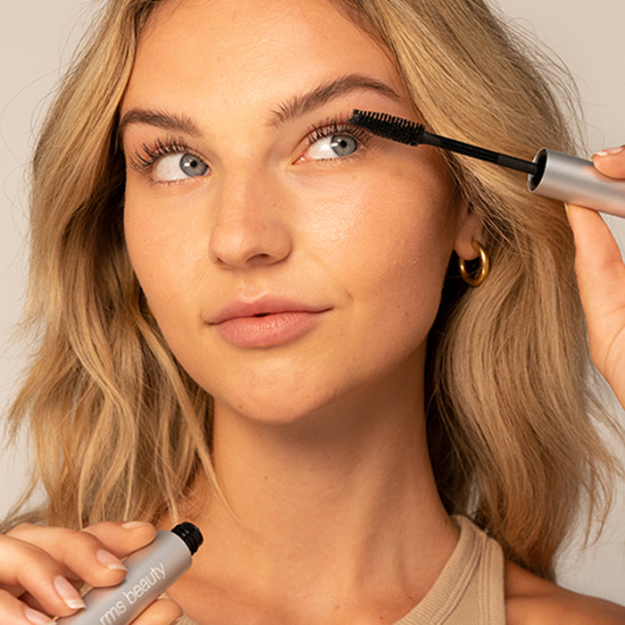A person with blonde hair uses RMS Straight Up™ Volumizing Peptide Mascara with a black wand, holding the tube in their other hand. They wear a tan sleeveless top and gold hoop earrings as peptides in the formula promise volume, complementing the neutral backdrop.