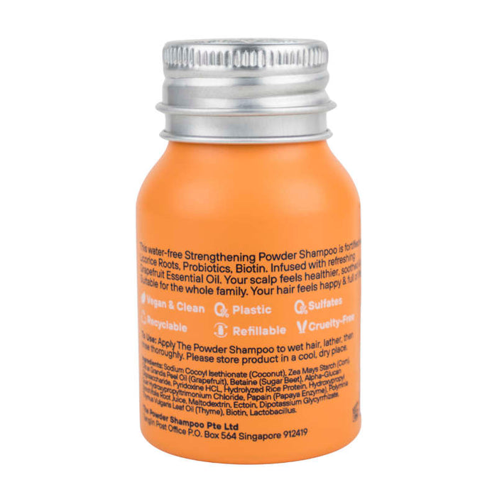 A small orange bottle of The Powder Shampoo’s Strengthening & Soothing Powder Shampoo features a silver cap, is vegan, cruelty-free, plastic-free, refillable, and infused with probiotics, biotin, and grapefruit essential oil for a botanical cleanse.