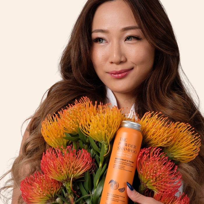 A person with long brown hair holds vibrant orange and yellow flowers, smiling gently. They present an orange bottle of Strengthening & Soothing Powder Shampoo by The Powder Shampoo, famous for its botanical ingredients that create a lush foam. The background is light beige.
