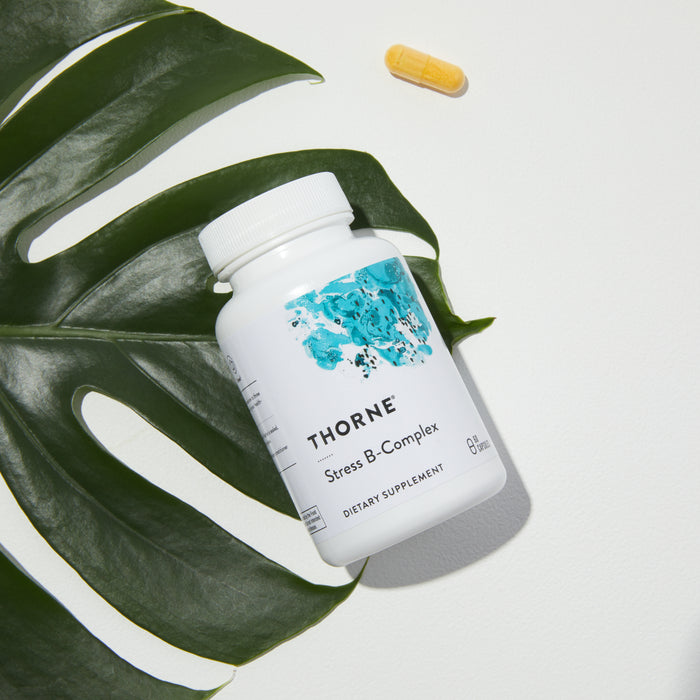 A white Thorne Stress B-Complex bottle, a dietary supplement for adrenal support rich in B vitamins, sits on a large green leaf with a single yellow capsule nearby on a white background.