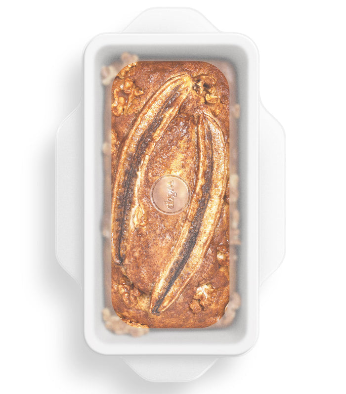 A rectangular white baking pan holds a loaf of banana bread topped with two banana halves. Viewed from above, this delightful creation stays fresh longer with W&P Stretch Baking Lids—an ideal food storage solution.