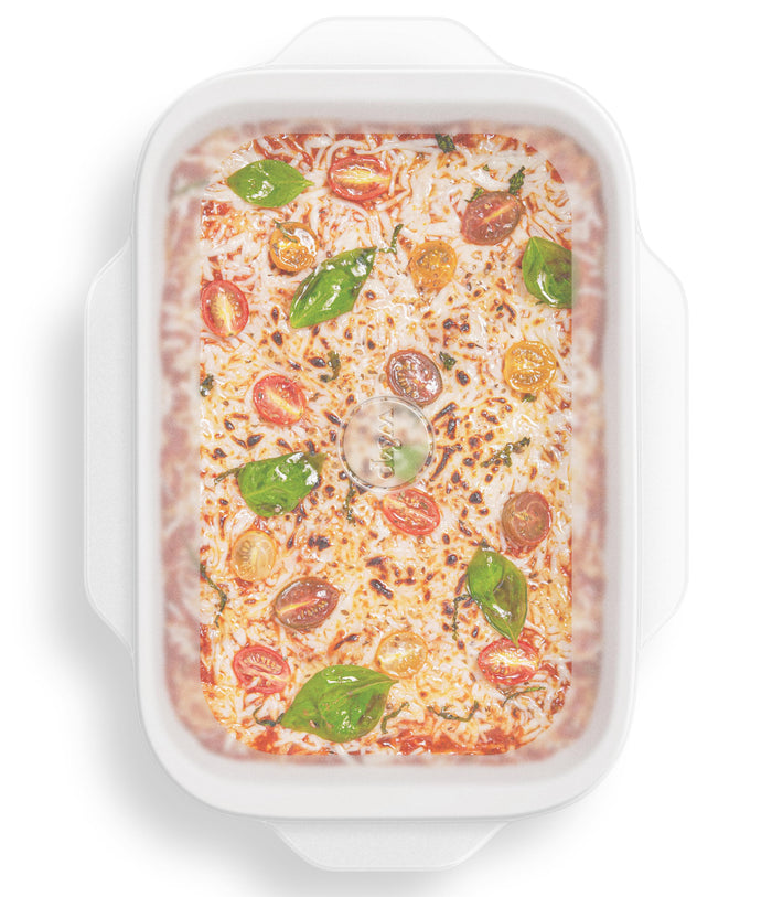 A top view of a rectangular white dish with baked pizza, melted cheese, halved cherry tomatoes, and basil leaves is shown alongside W&Ps Stretch Baking Lids. The dish has side handles for easy use, and the lids are perfect for effortless food storage solutions.