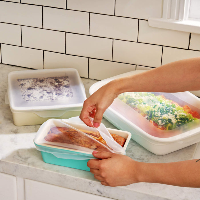 A person seals a reusable green food storage container with cooked chicken inside. Other containers, featuring W&Ps Stretch Baking Lids, hold a cooked meal, vegetables, and a frozen item on the marble countertop. A white tiled wall in the background highlights smart food storage solutions.