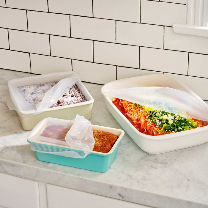Three silicone dishes with W&Ps Stretch Baking Lids sit on a marble countertop. One holds a red and green vegetable bake, another a casserole, and the third a loaf of bread, all staying fresh against the white tiled wall backdrop with these innovative food storage solutions.
