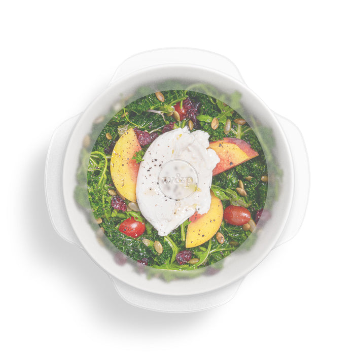 Savor a fresh salad of leafy greens, cherry tomatoes, peach slices, pepitas, and a poached egg in a clear bowl with a white lid. W&Ps Stretch Lids provide the perfect storage solution to maintain freshness every time you open it.