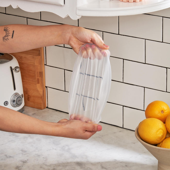In the kitchen, someone uses a W&P Stretch Lid to cover a small plate. The scene includes white subway tiles, a wooden block, and a bowl of lemons on the marble countertop.