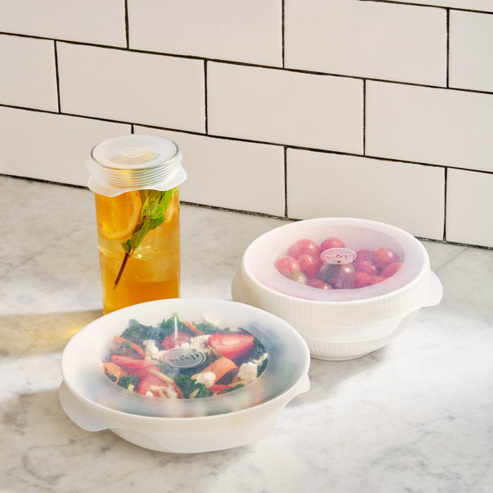 Stretch Lids from W&P securely cover containers on a marble countertop. One container holds iced tea with lemon and mint, another has a fresh salad with vegetables, and the third is filled with grapes. The storage setup is set against a tiled wall in the background.