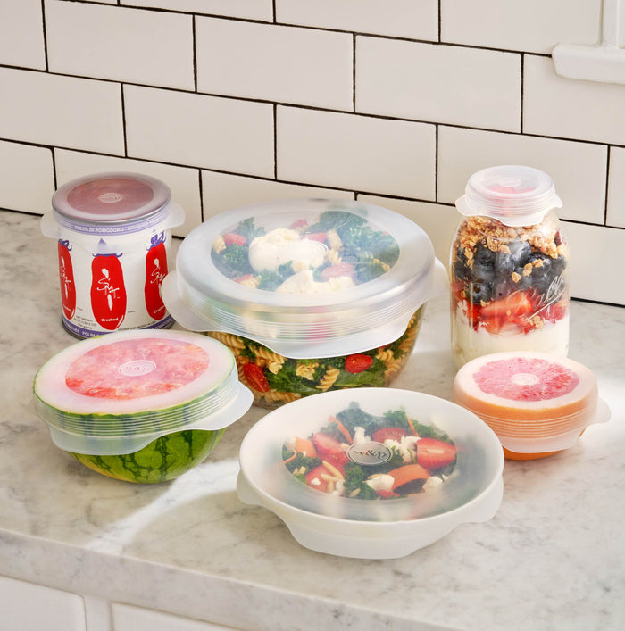 A kitchen countertop features various food containers, each sealed with W&P Stretch Lids, for a smart storage solution. These translucent reusable lids cover salad, yogurt with berries, and sliced fruit against a tiled backsplash.
