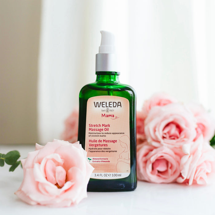 A green Weleda Stretch Mark Massage Oil bottle with a pump sits on a white surface, surrounded by pink roses and soft white curtains in the background. Perfect for pregnancy skincare, the label highlights its benefits for reducing stretch marks.